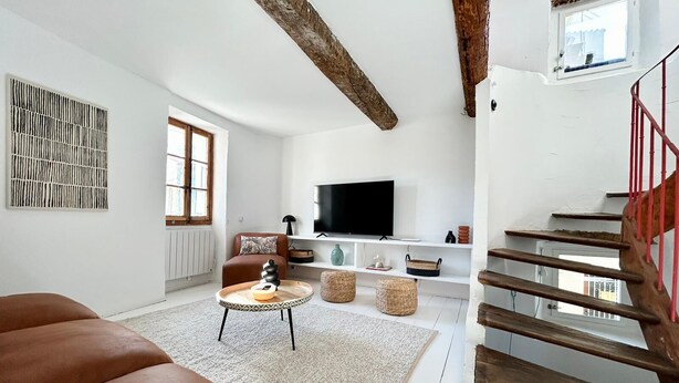 A spacious living room with a large brown sofa, a wooden staircase with red railings, a television on a white shelf, and a window with wooden beams on the ceiling. - Atypical sunny house - Panoramic terrace at the foot of the Albères (Sorède)