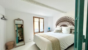 A bedroom with a large bed, a wooden headboard, a mirror, a window with curtains, and a small stool. - Atypical sunny house - Panoramic terrace at the foot of the Albères (Sorède)