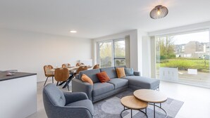 Un salon moderne avec un canapé gris, un coin repas comprenant une table et des chaises, et de grandes fenêtres offrant une vue sur l’extérieur.