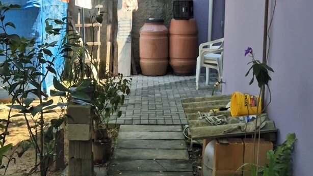 En smal sti, der fører hen til en bygning med lilla facade, omgivet af plantekasser og udendørs møbler.