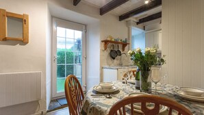 Un comedor con una mesa dispuesta para cuatro personas, sillas, un jarrón con flores y una zona de cocina con utensilios colgados en la pared.