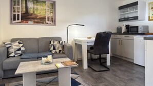 Een moderne, compacte leefruimte met een grijze bank, een klein tafeltje met een boek en kaarsen, een bureau met een lamp, een keuken met kasten en een magnetron, en een groot raam met uitzicht op een bos.