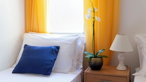 A bed with white linens and a blue pillow, a bedside table with a lamp and a potted plant, and a window with yellow curtains.