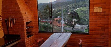 A rustic dining area with a large wooden table, two hanging light bulbs, and a view of a hilly landscape through a large window.