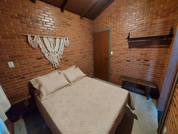 Une pièce avec un mur en briques, un lit recouvert d’une courtepointe beige, une porte en bois et une petite table.