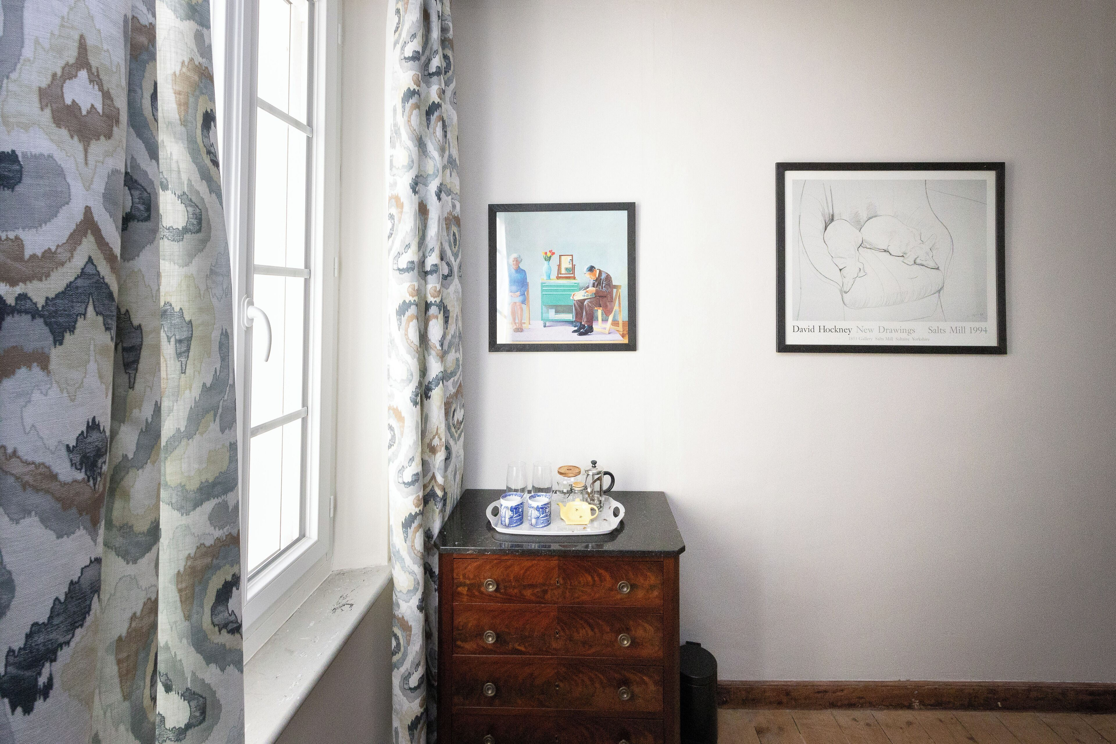 A room with a window, a wooden dresser with a tray of items, and two framed artworks on the wall.
