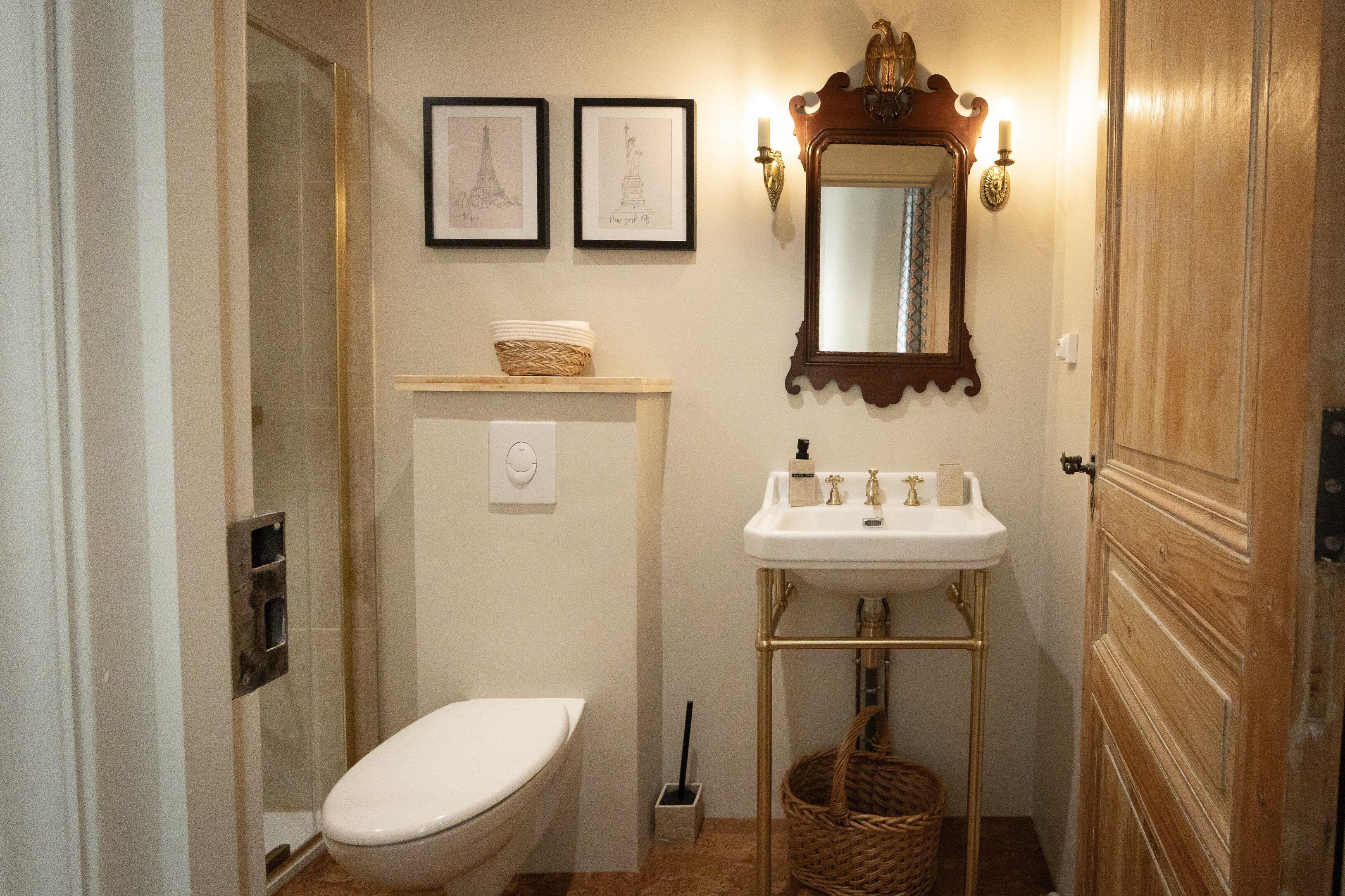 A bathroom with a toilet, sink, and shower. There are two framed pictures on the wall, a mirror above the sink, and a wooden door.