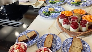 Eine Küche mit Herd, Töpfen und einer Person beim Kochen. Es gibt Teller mit Essen, einen Brotkorb sowie Schüsseln mit Salat und Obst.