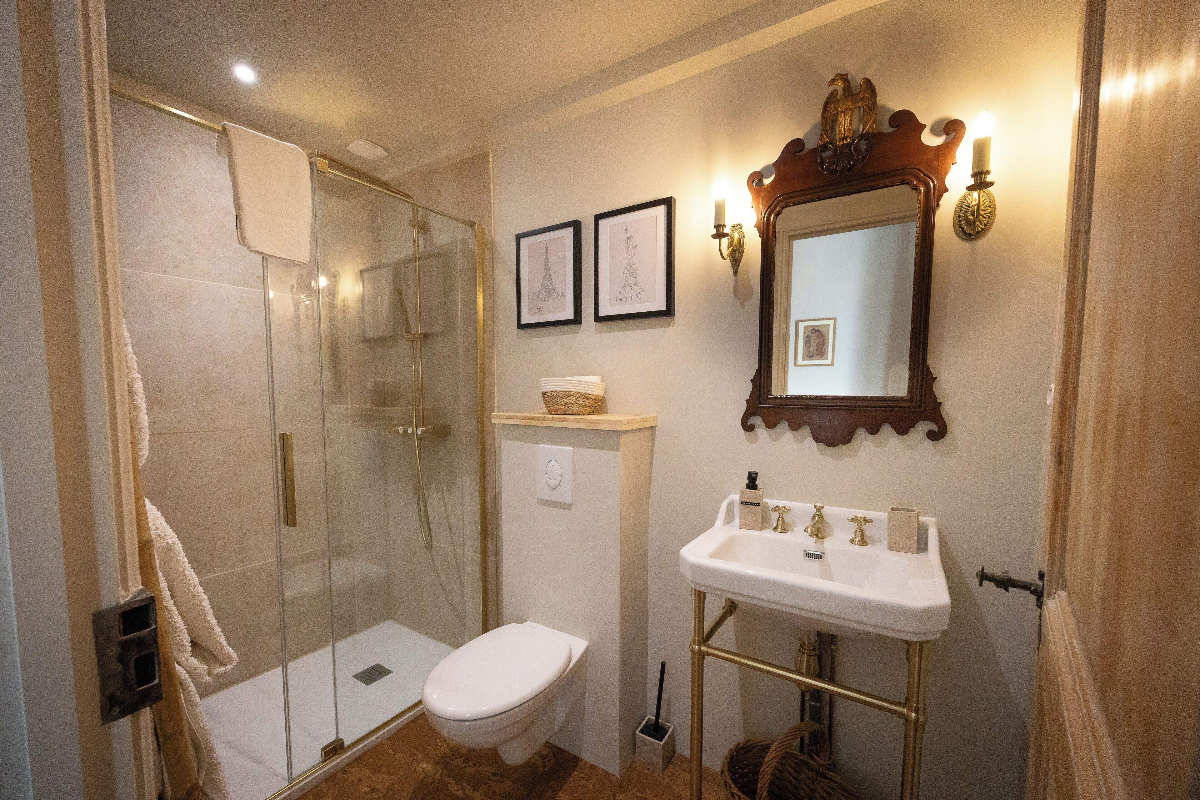 A bathroom with a glass-enclosed shower, a white toilet, a sink with a gold pedestal, and framed pictures on the wall.