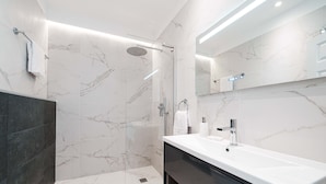 Un baño moderno con ducha de vidrio, un lavamanos blanco y un mueble oscuro, y paredes de mármol.