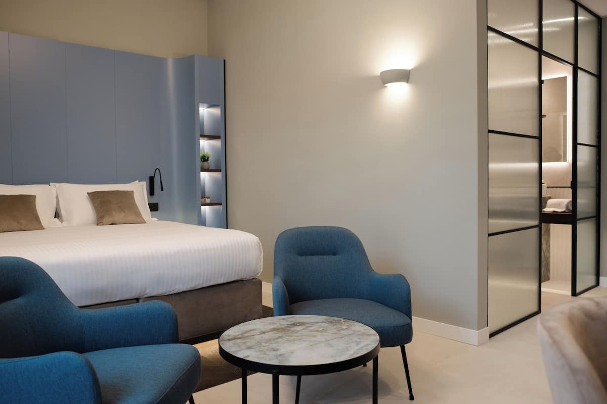A modern hotel room with a bed, two blue armchairs, a small round table, and a built-in wardrobe with a sliding door.