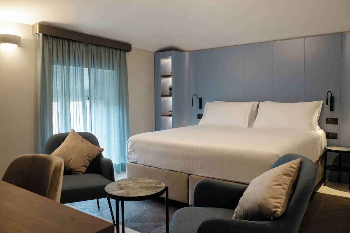 A modern hotel room with a large bed, two armchairs, a small table, and a built-in shelf with decorative items.