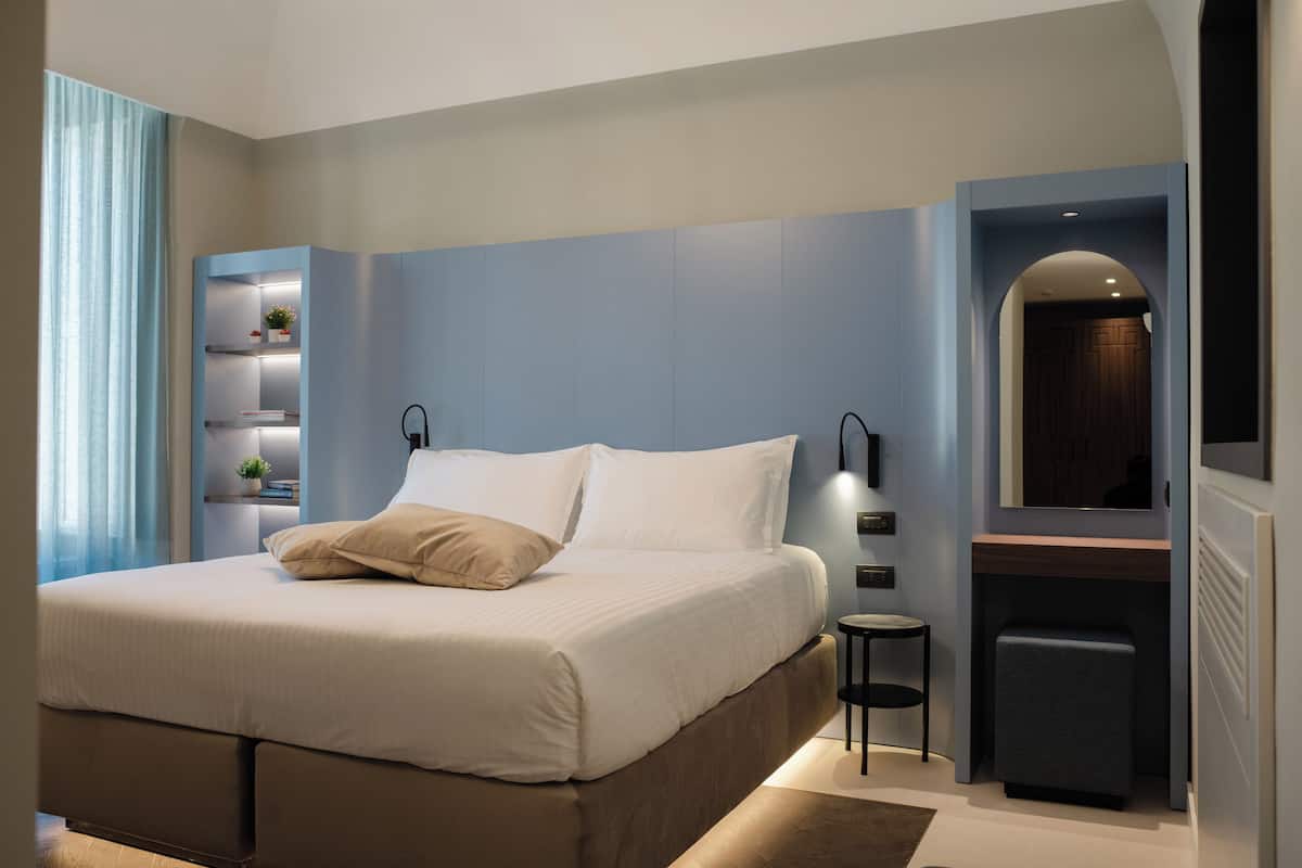 A modern bedroom with a large bed, bedside tables, a built-in shelf with plants, and a mirror above the door.