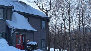 Een huis met twee verdiepingen, met een rode deur en ramen, omringd door besneeuwde grond en bomen.