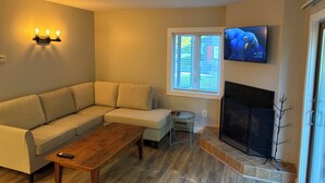 Een woonkamer met een hoekbank, een houten salontafel, een open haard en een aan de muur bevestigde televisie.