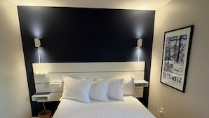 Een net opgemaakt bed met een witte dekbedovertrek en kussens, een donker hoofdbord, nachtkastjes met lampen en een ingelijste foto aan de muur.