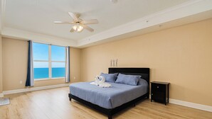 Quarto com uma cama grande, um ventilador de teto, mesa de cabeceira e vista para o oceano através de uma janela ampla.