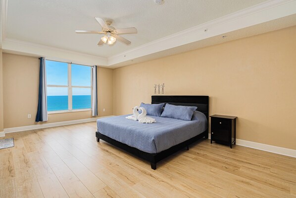 A bedroom with a large bed, a ceiling fan, a nightstand, and a view of the ocean through a large window. - Oceanfront | Heated Indoor/Outdoor Pool-St. Maarten #806 (Daytona Beach Shores)