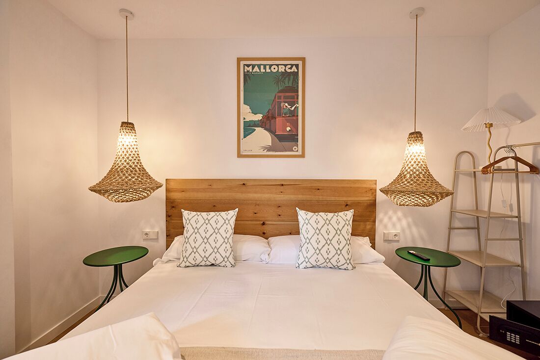 A hotel room with a wooden headboard, two hanging lamps, a framed picture of Mallorca, a bed with patterned pillows, a green side table, and a small shelf with a remote control.