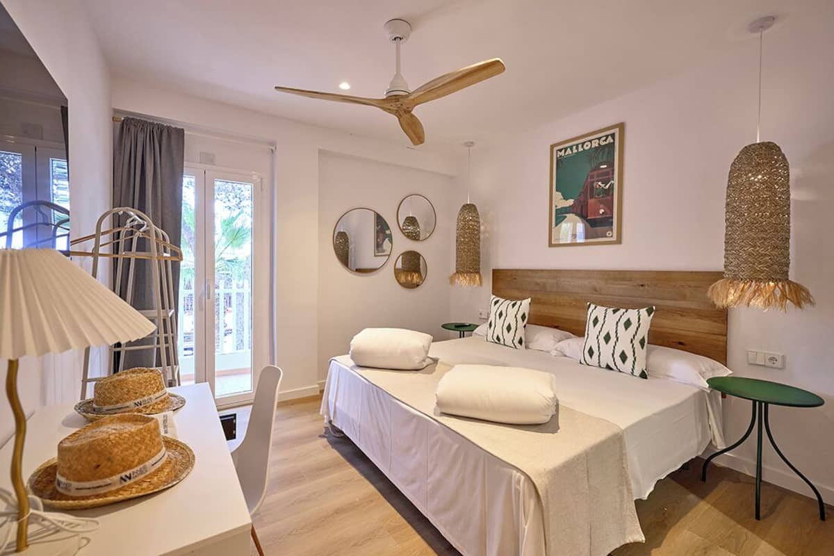 A hotel room with a bed, a ceiling fan, a table with hats, a chair, and a poster on the wall.