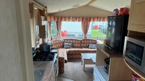 Une cuisine compacte pour camping-car, équipée d’une cuisinière, d’un four à micro-ondes et d’un réfrigérateur. Un canapé est placé au centre, et un téléviseur est fixé au mur au-dessus.
