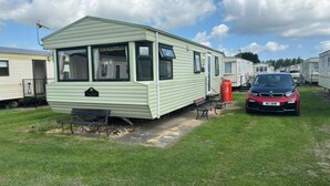 Une maison mobile avec un porche à l’avant, un banc et une bouteille de gaz rouge.