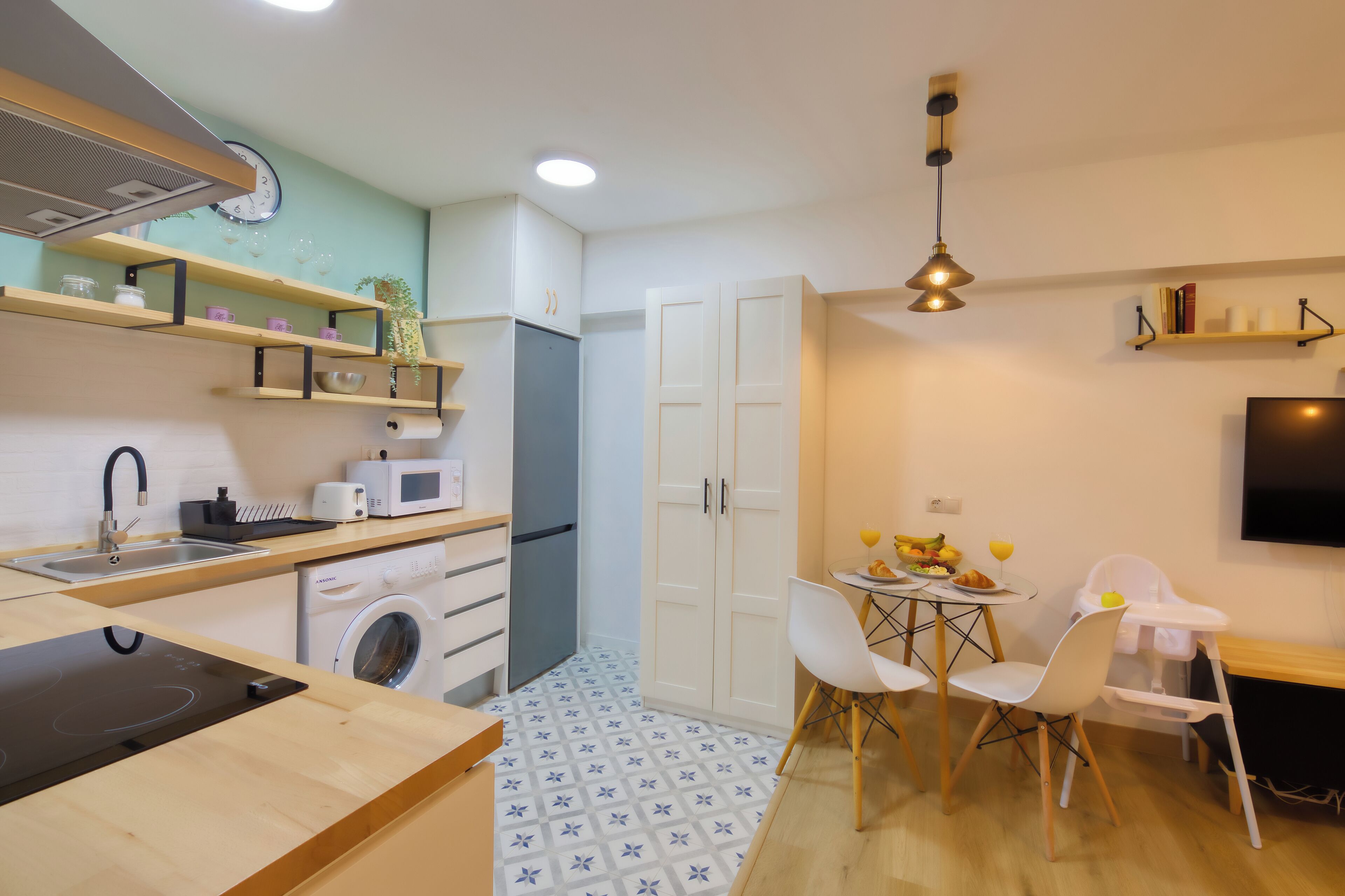 A compact kitchen with a microwave, sink, and washing machine. There is a dining area with a table, chairs, and a baby high chair.
