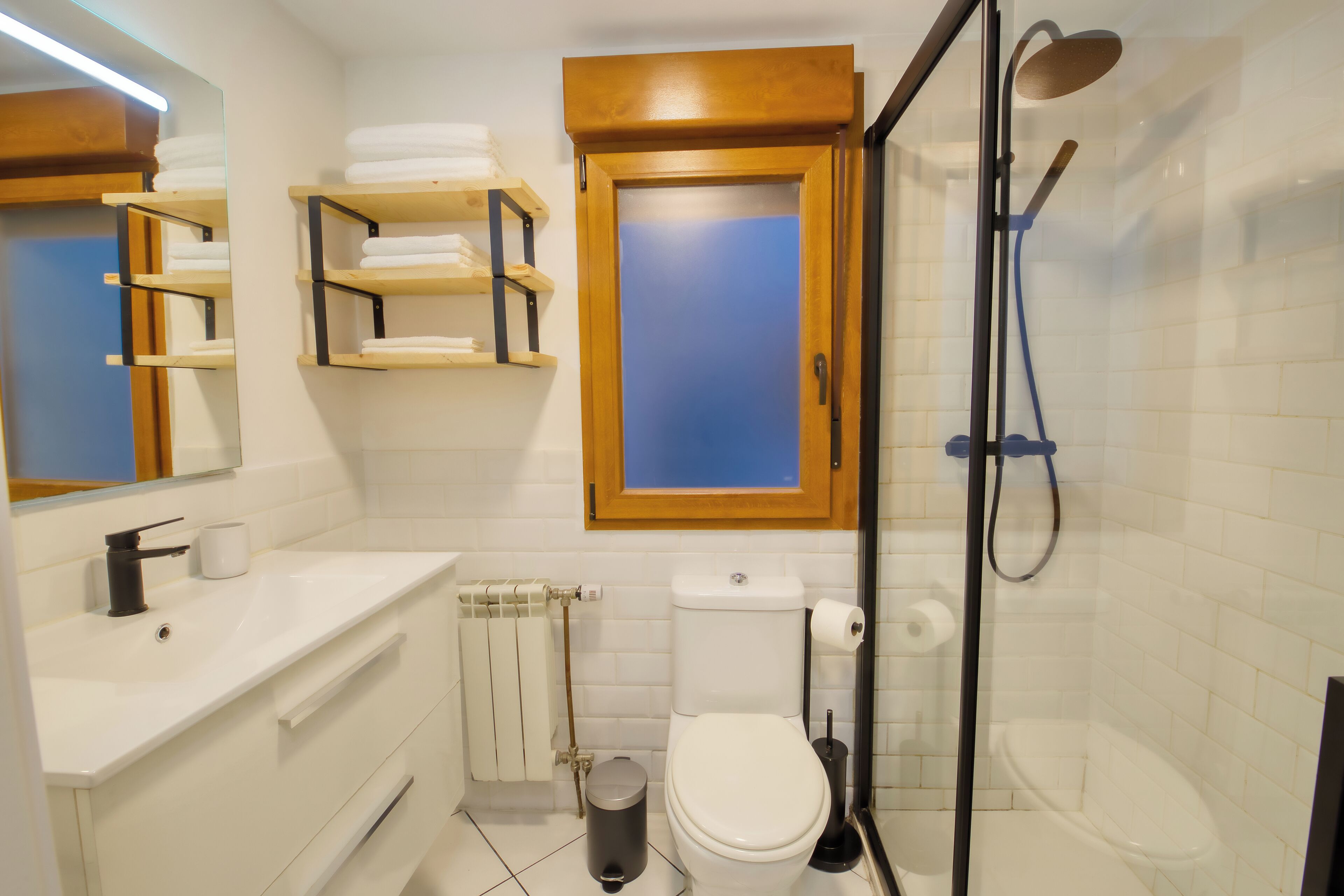 Un baño con lavamanos, inodoro y ducha blancos. Hay repisas sobre el lavamanos y una puerta de madera sobre el inodoro.