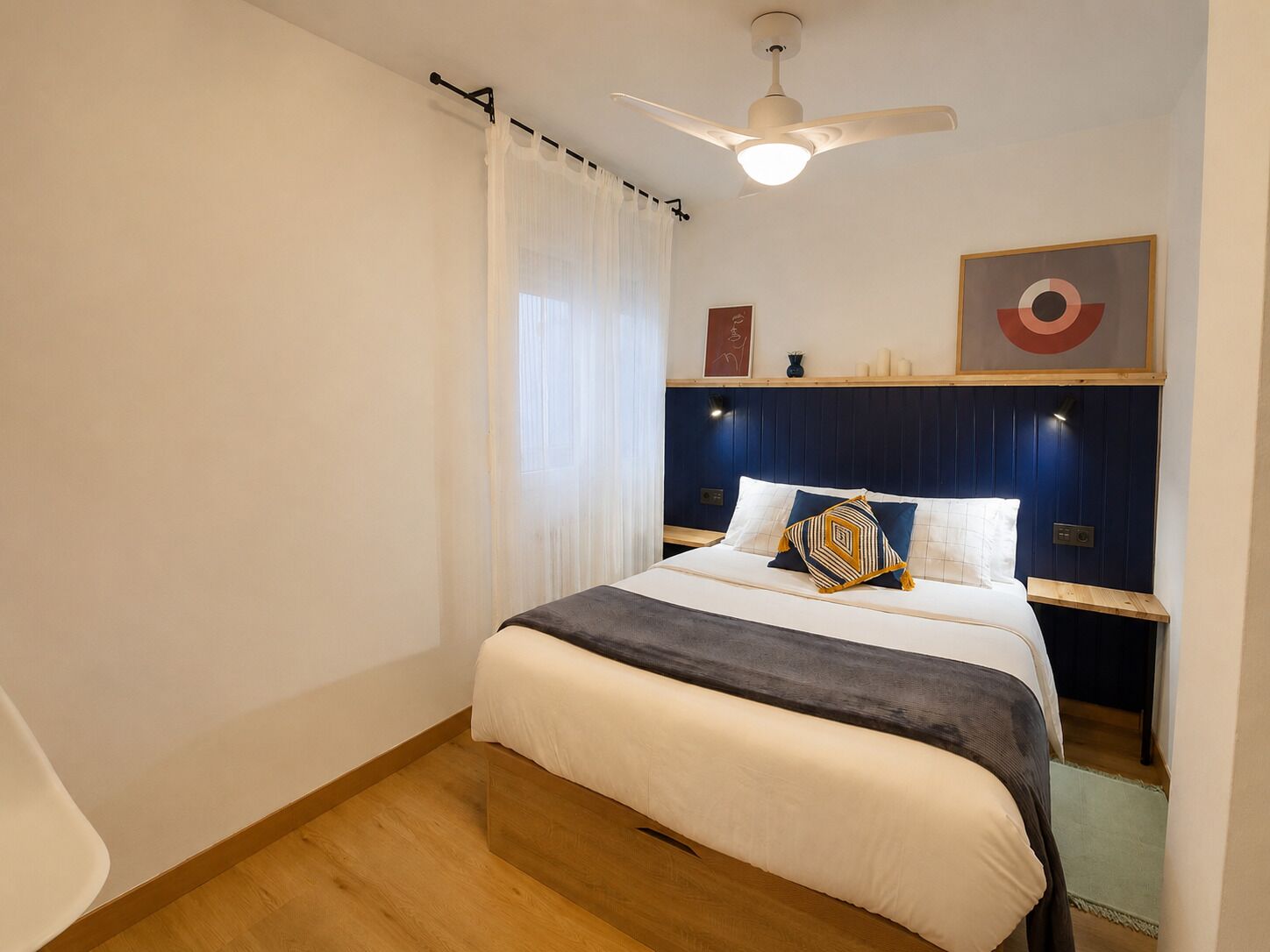 A hotel room with a bed, a ceiling fan, a nightstand, a wall-mounted shelf, and a window with curtains.
