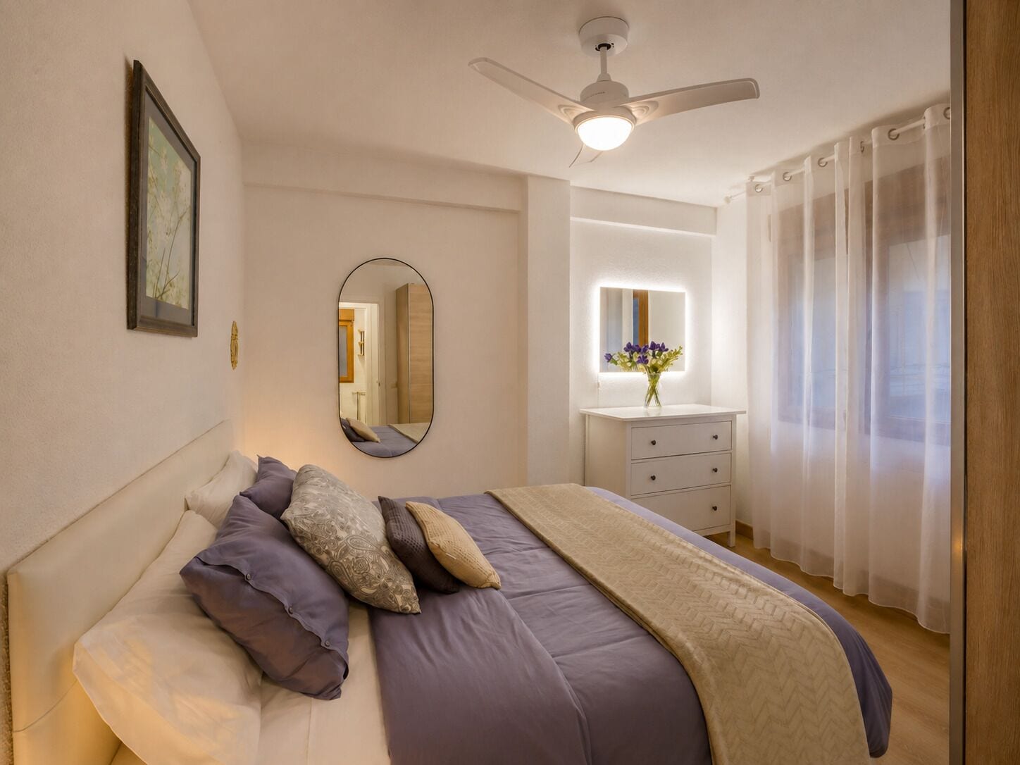 A bedroom with a bed, a ceiling fan, a mirror, a dresser, and a window with curtains.