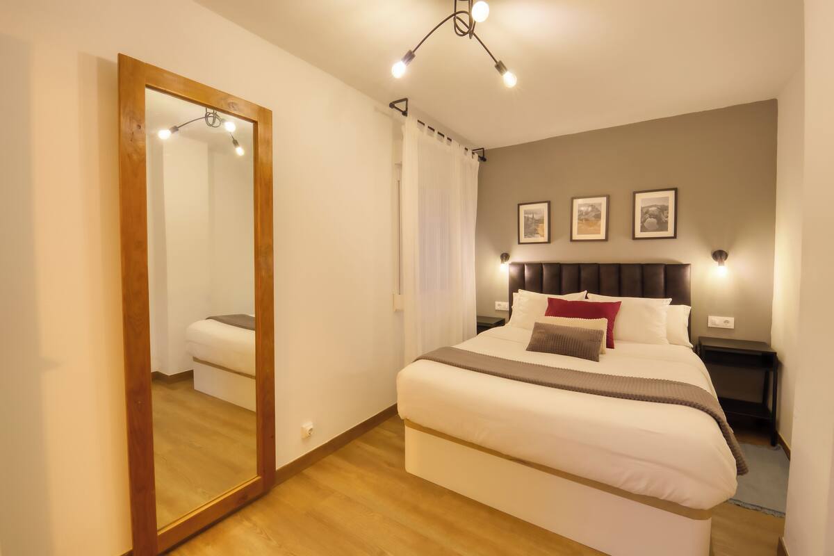 A bedroom with a large mirror, a bed with white bedding, a headboard with lights, and two framed pictures on the wall.
