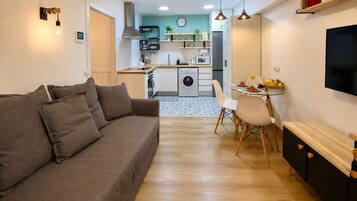 Un espace de vie compact comprenant un canapé, une table à manger, des chaises, un téléviseur, un coin cuisine avec un lave-linge et une horloge au mur.