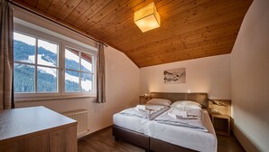 A bedroom with a wooden ceiling, a large window with a view of snow-covered mountains, a bed with white linens, and a wooden desk.