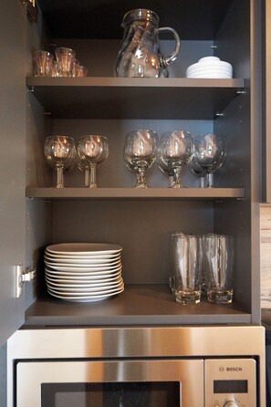 Una cocina con un microondas Bosch, estantes con vasos, platos y una jarra.