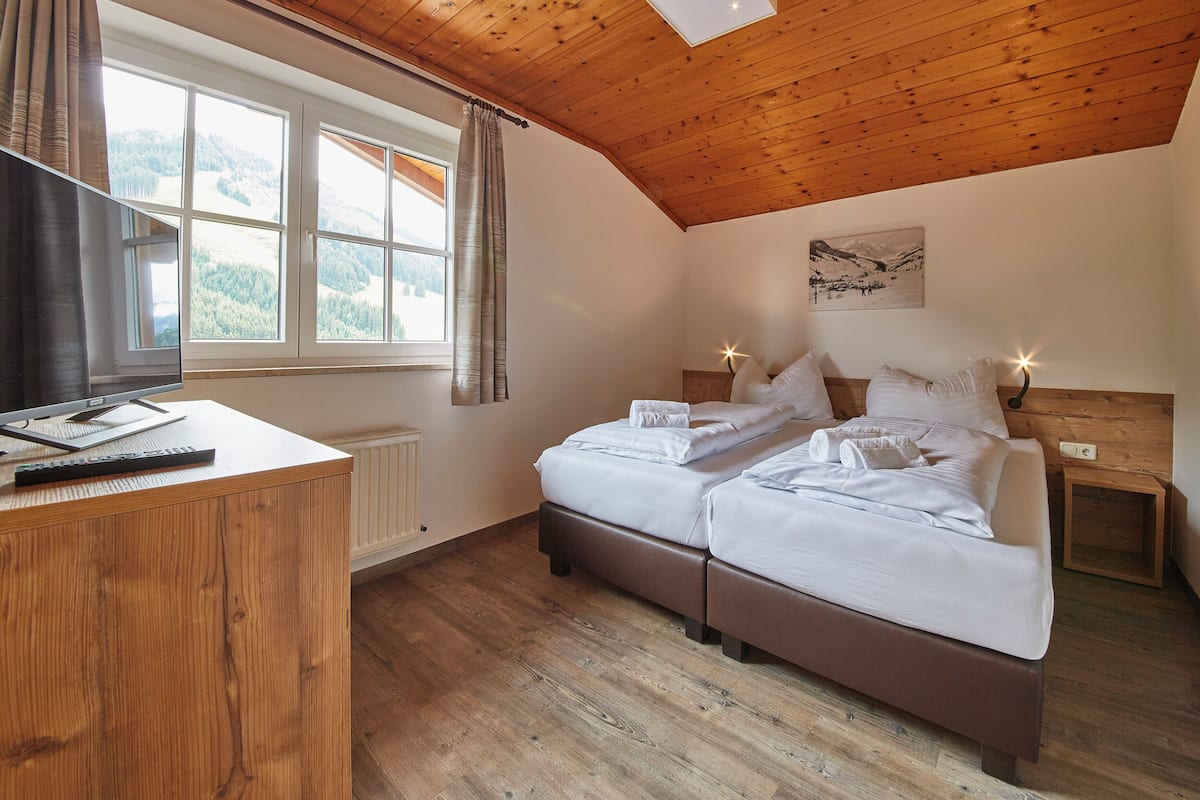 A bedroom with two beds, a wooden ceiling, a window with curtains, a radiator, and a painting on the wall.