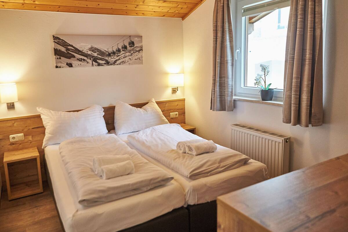 A double bed with white linens, two bedside tables, a wall-mounted light, a window with curtains, and a black and white photo on the wall.