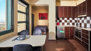 A compact hotel room with a kitchenette, a bed, a small table, and a view of the city through the window.