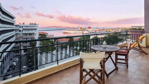 A balcony with a table and chairs, overlooking a waterfront with a cruise ship and cityscape.