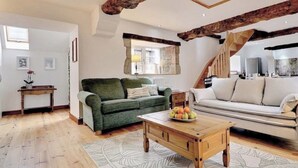 Un salon spacieux avec une table basse en bois, deux canapés, une fenêtre encadrée de pierres et des poutres apparentes.