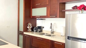 A modern kitchen with wooden cabinets, a stainless steel refrigerator, and a countertop with a coffee maker and cups.
