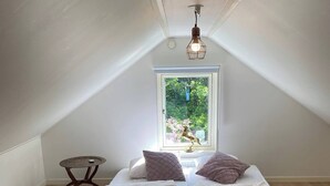Una piccola e accogliente camera da letto in mansarda, con soffitto spiovente, una finestra vista su del verde, un letto con due cuscini e un tavolino.