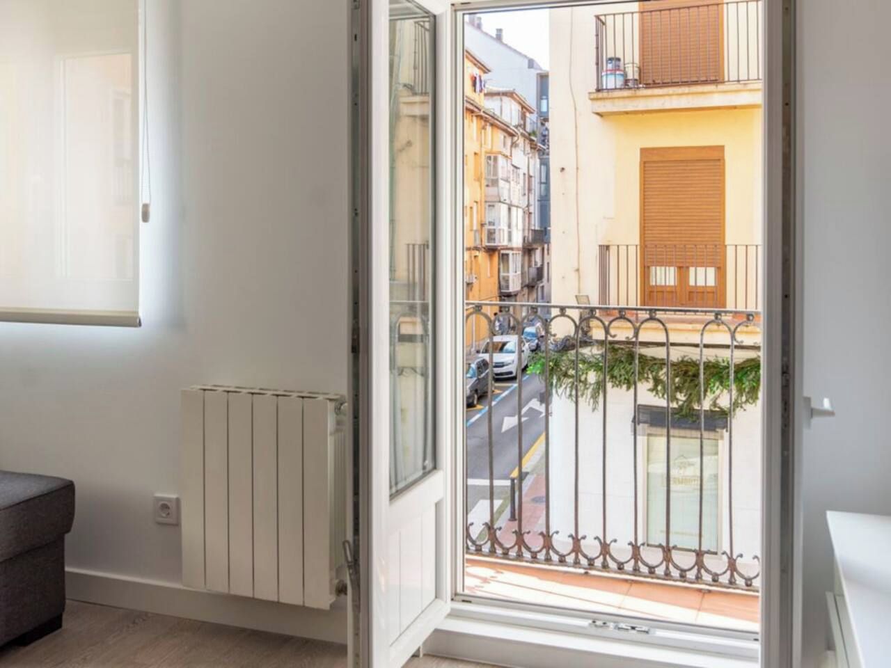 Una habitación con un radiador, una puerta que da a un balcón con barandilla metálica y vistas a una calle con edificios.