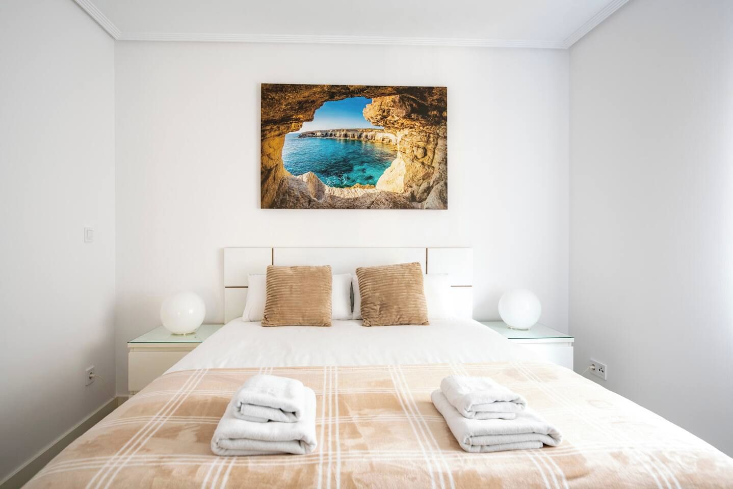 Habitación de hotel con cama, dos almohadas, mesita de noche, lámpara y un cuadro de una escena costera.