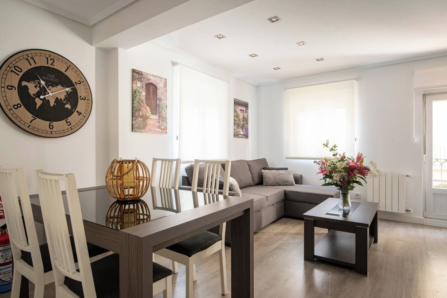 A modern living room with a large wall clock, a sofa, a dining table, and a window with natural light.