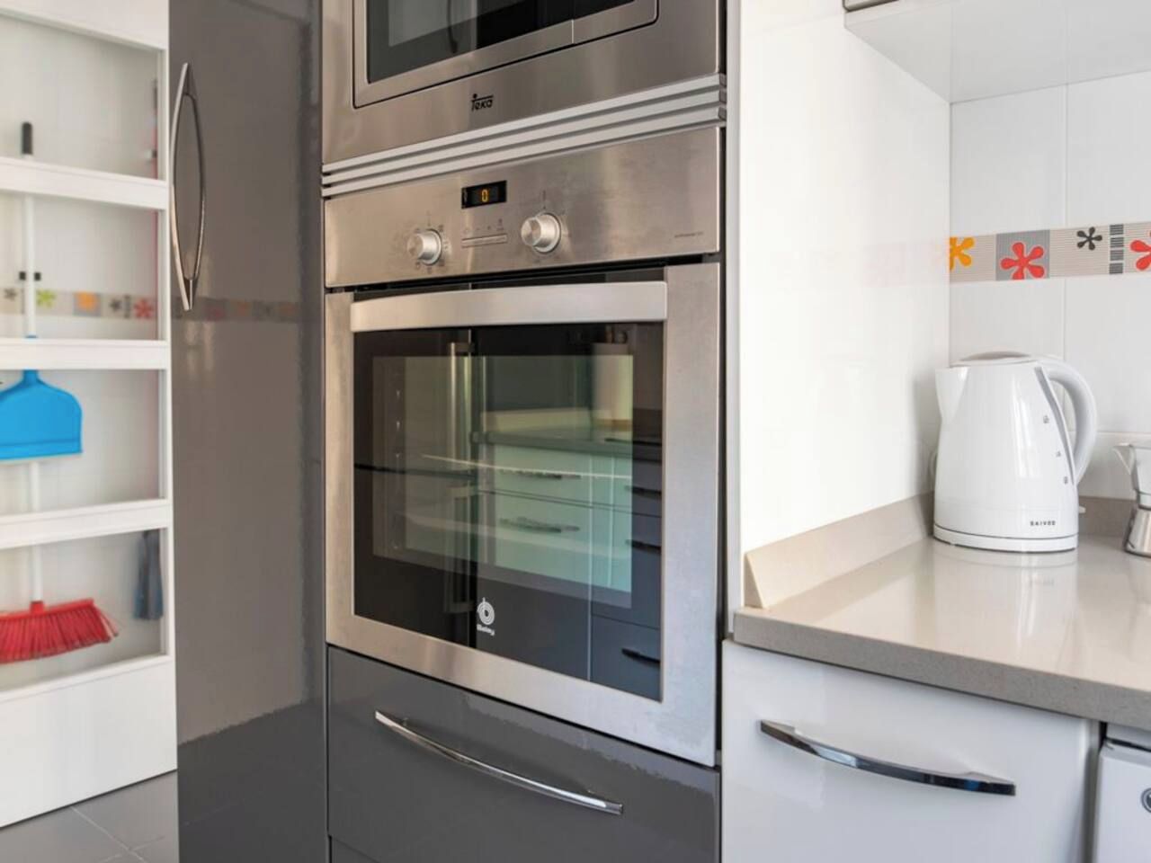 A kitchen with a built-in oven and microwave, a kettle, and a shelf with cleaning supplies.
