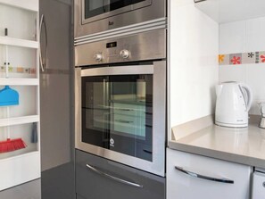 Una cocina con horno empotrado y microondas, hervidor y estante con productos de limpieza.