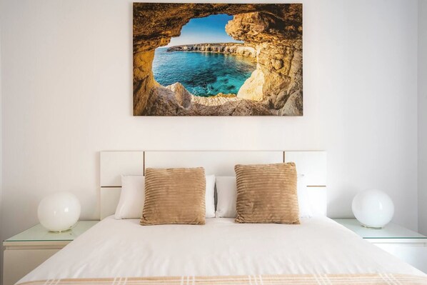 A bed with white bedding and two beige pillows, a white headboard, and a wall-mounted painting of a coastal landscape.