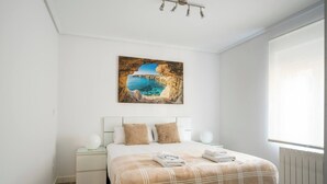 A bedroom with a bed, a nightstand, a wall-mounted lamp, a painting of a coastal scene, and a window with blinds. - Apartamento Atalaya en el centro (Santander)