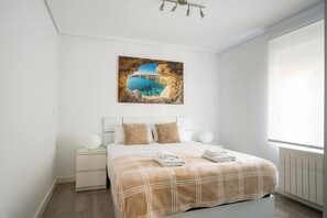 Un dormitorio con cama, mesita de noche, lámpara de pared, cuadro de una escena costera y ventana con persianas.