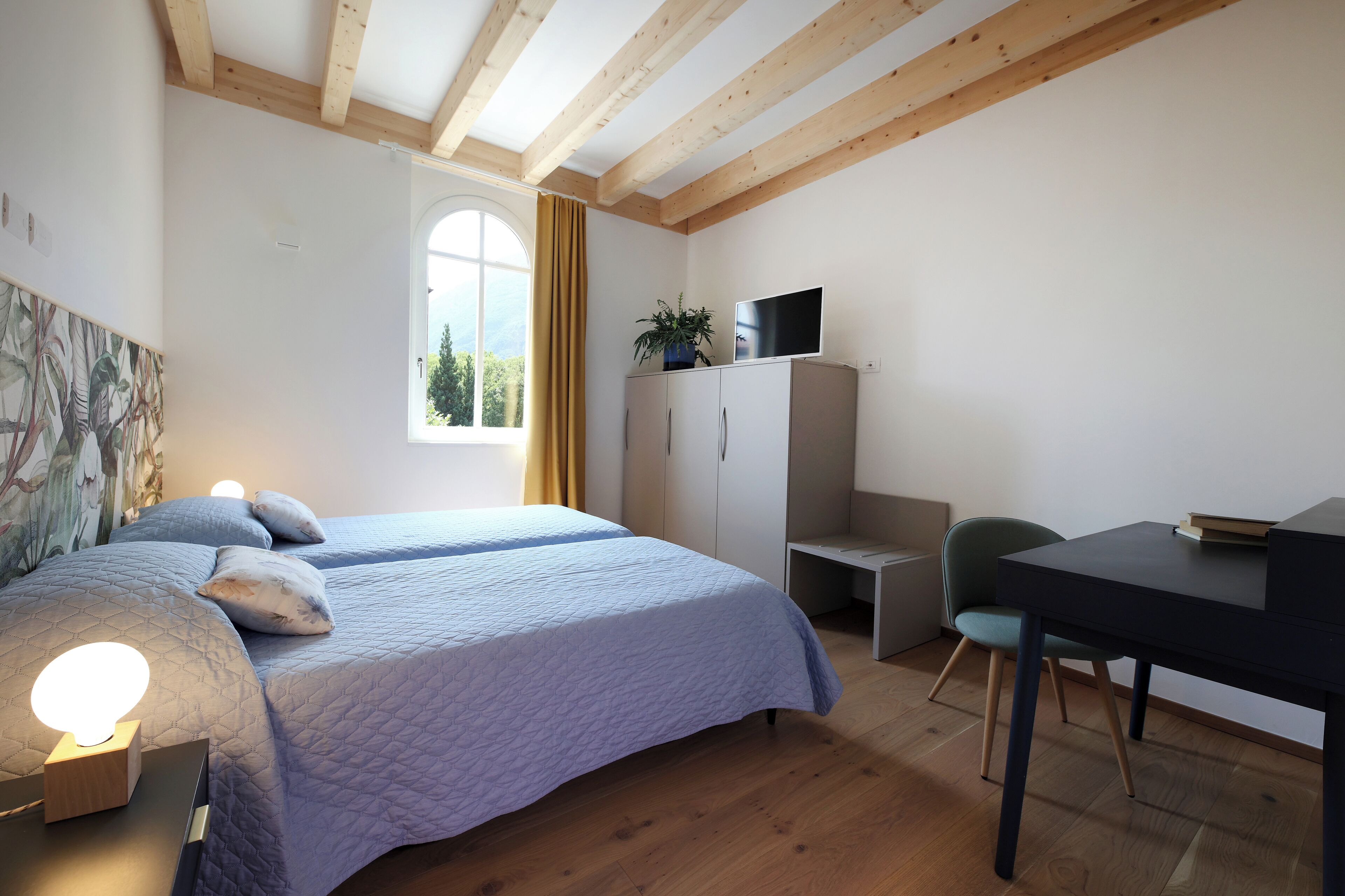 A bedroom with a large bed, a desk, a chair, a TV, and a window with curtains.